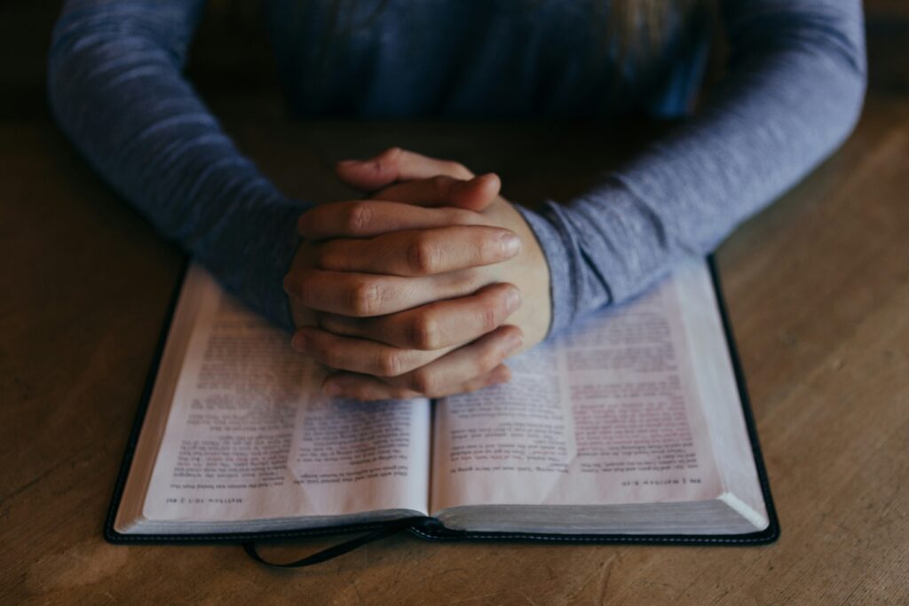 Person praying with hands folded over open Bible on wooden table