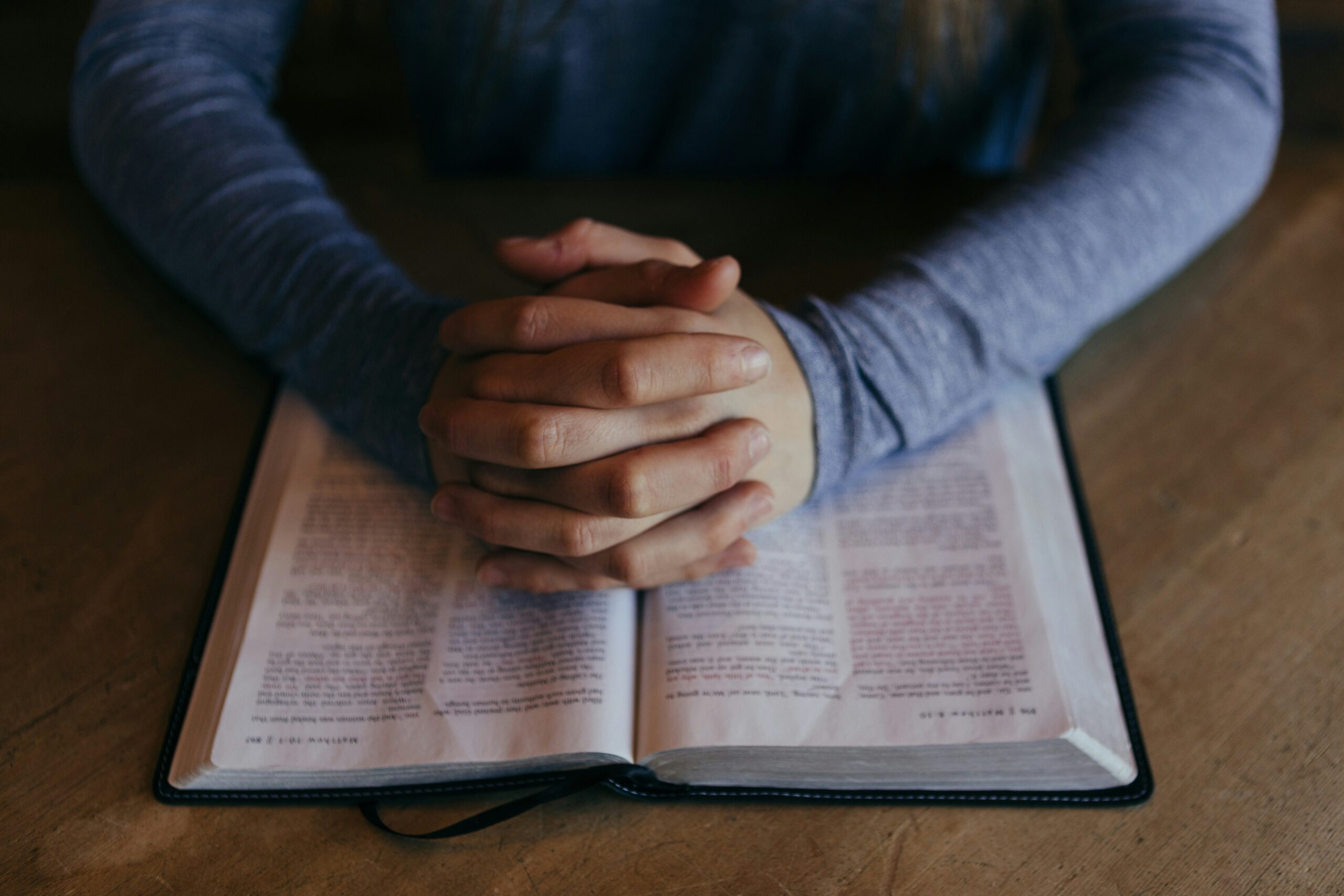 Person praying with hands folded over open Bible on wooden table