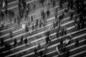 Blurry black and white photo of a busy crowd crossing a street, symbolizing the rush of modern life